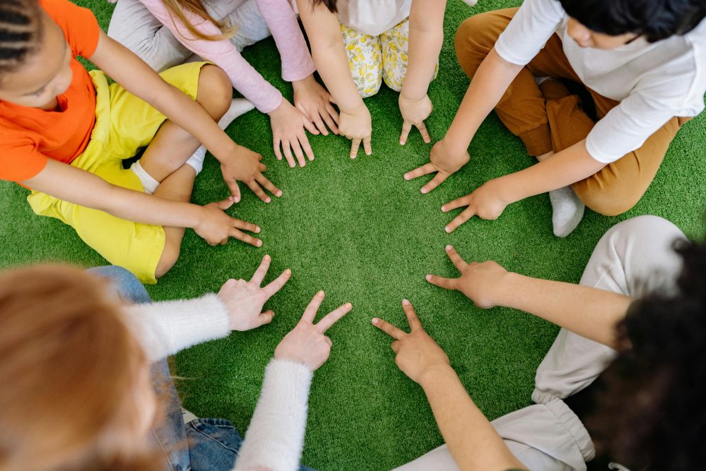 pexels-pavel-danilyuk-8422173 Children sitting in a circle on the floor leaning forward with hands out and fingers holding the peace sign down to the green carpet
